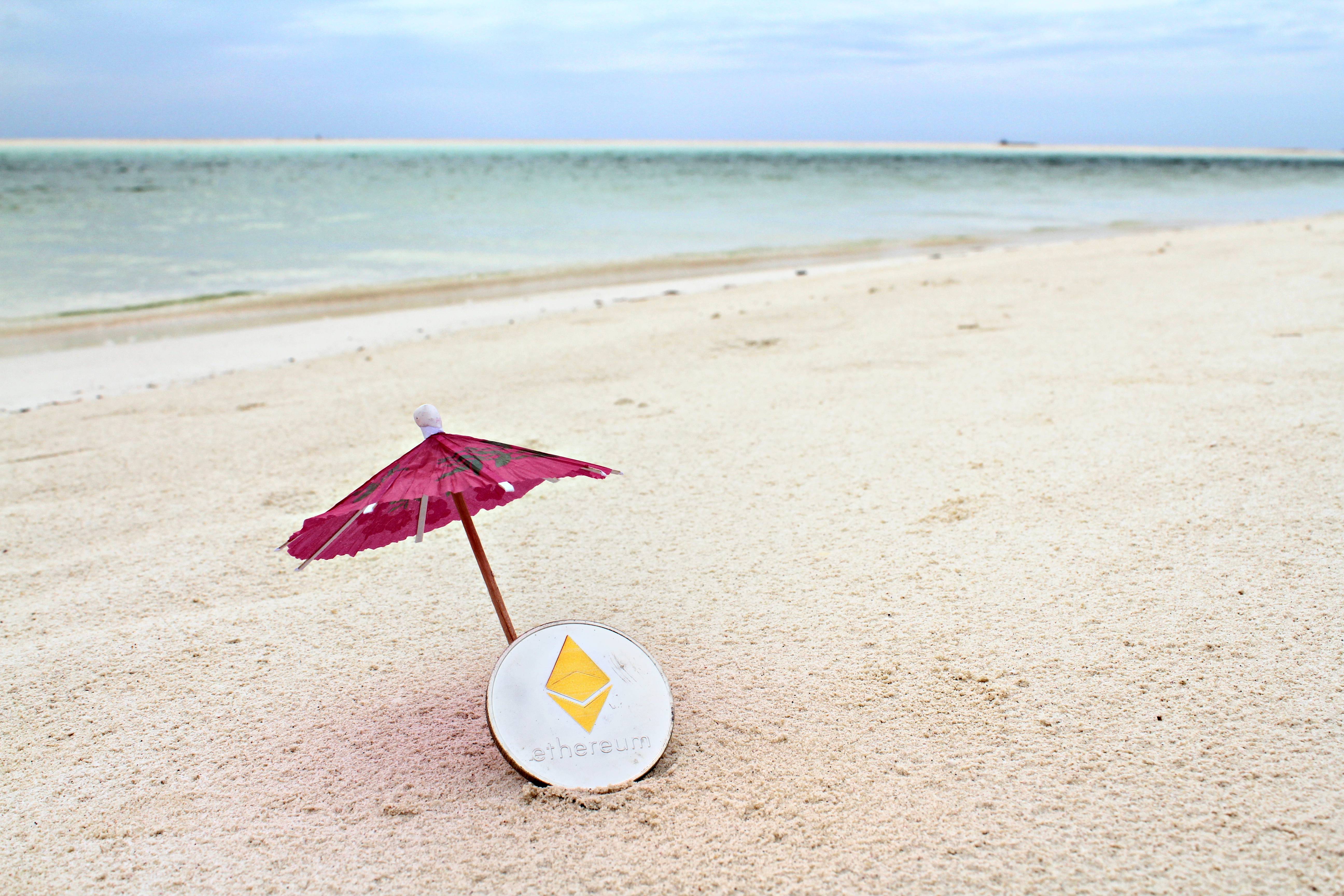 Silver Ethereum coin under the paper umbrella on the beach, shal